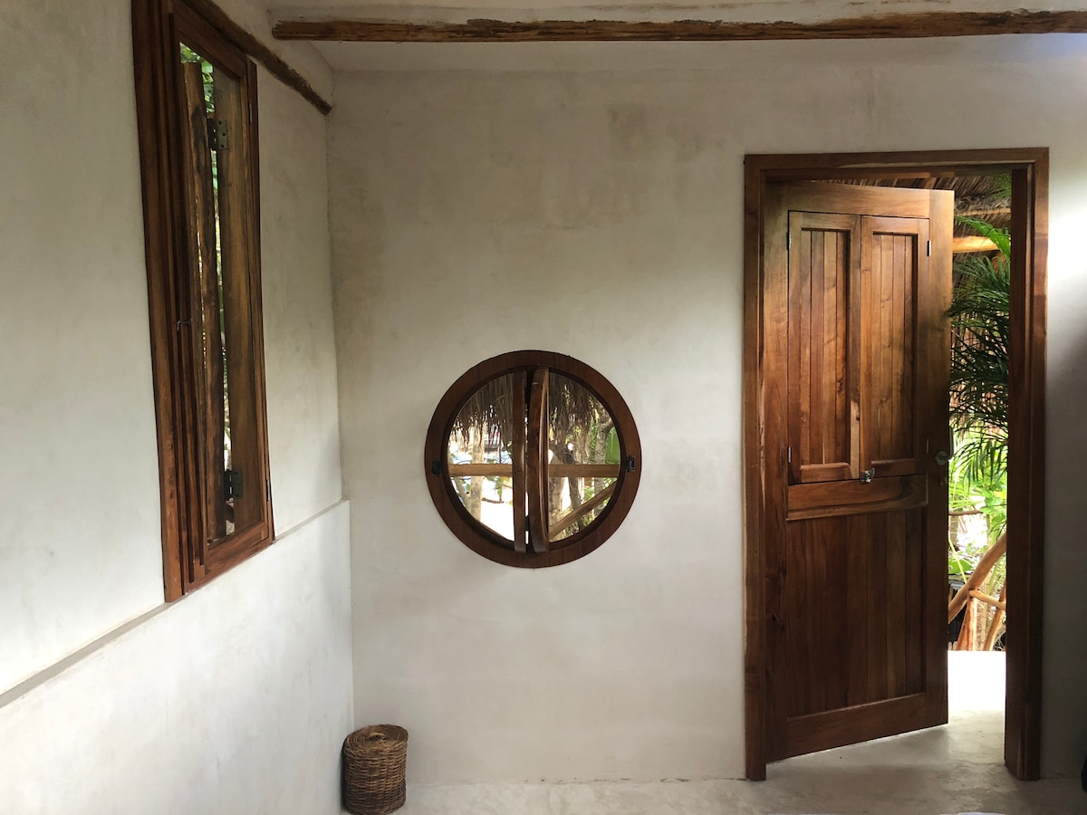 A simple entrance area features a round window providing a view of the lush outdoors. The wooden door stands ajar, allowing natural light to enter. Soft, neutral walls are complemented by natural wood accents, creating a calm and welcoming entryway.