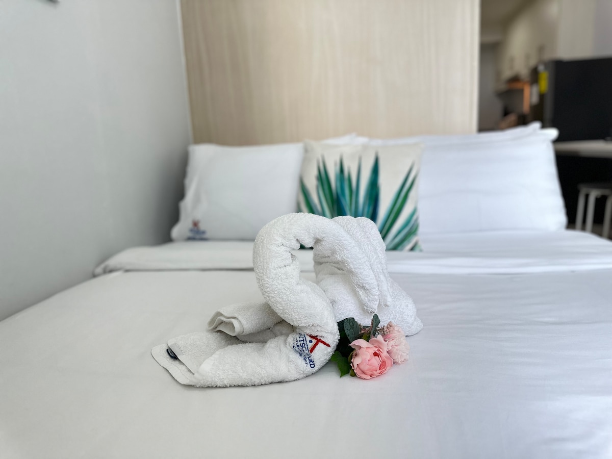 A neatly made bed is presented with white linens and decorative cushions. A towel is elegantly folded into a decorative shape on the bed, accompanied by a small artificial flower. The soft, neutral tones create a calming setting.