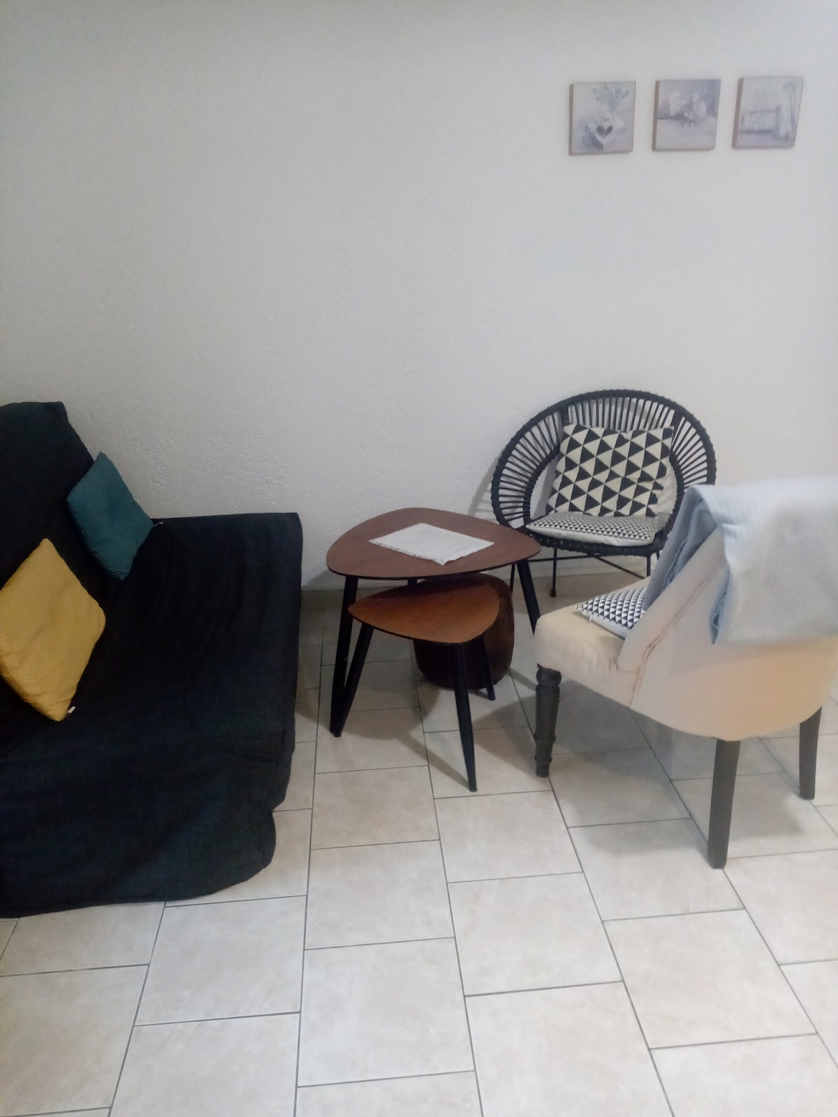 A cozy seating area is displayed, featuring a dark sofa adorned with colorful cushions and a patterned chair. A small, triangular coffee table is positioned in between. Light-colored tiles cover the floor, and three framed pictures hang on the wall above the seating.