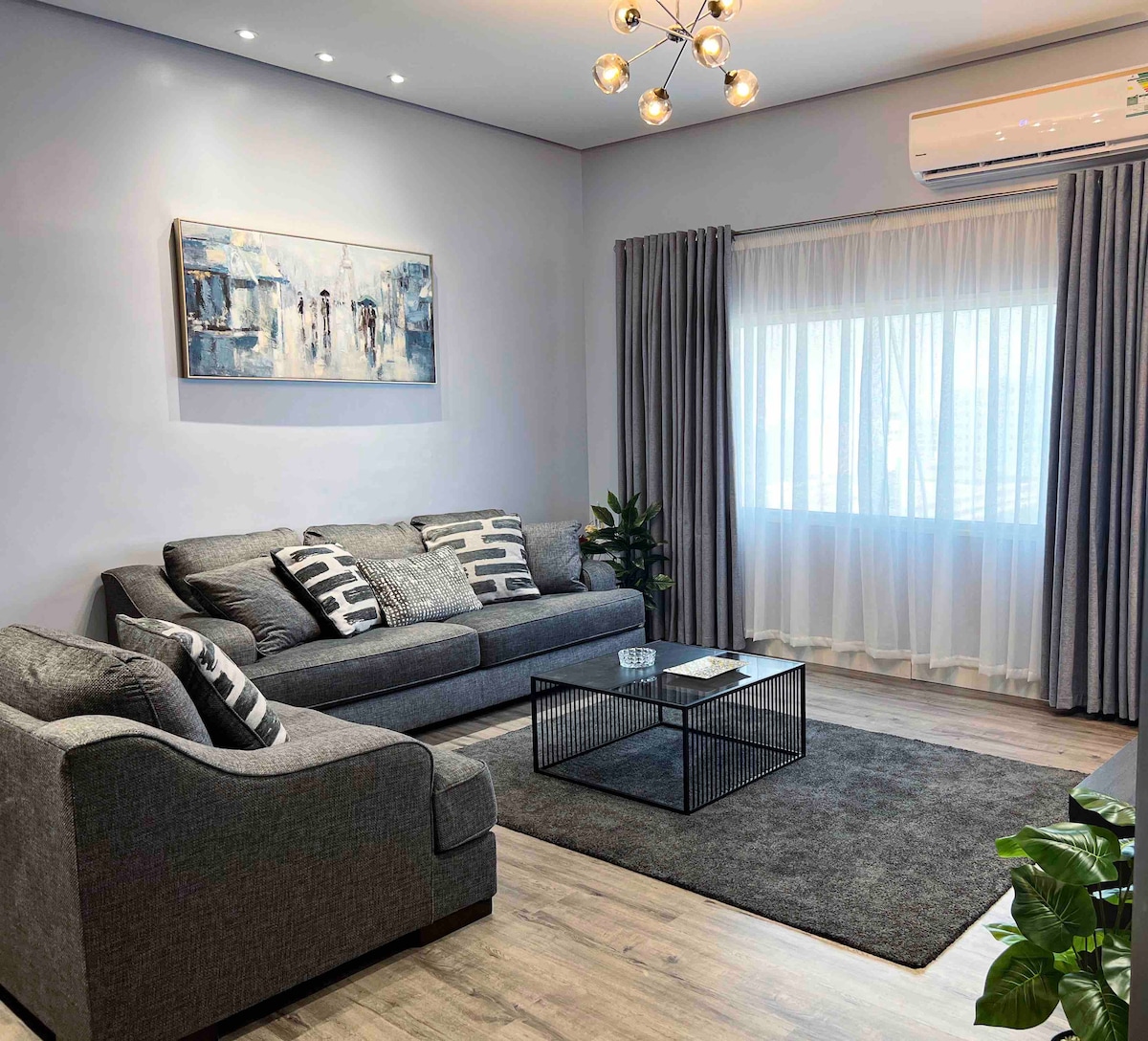 A modern living area is presented with a comfortable grey sofa and armchair, adorned with patterned cushions. A sleek black coffee table sits atop a soft area rug, and a large window with sheer curtains allows natural light to brighten the space, creating a relaxed environment.