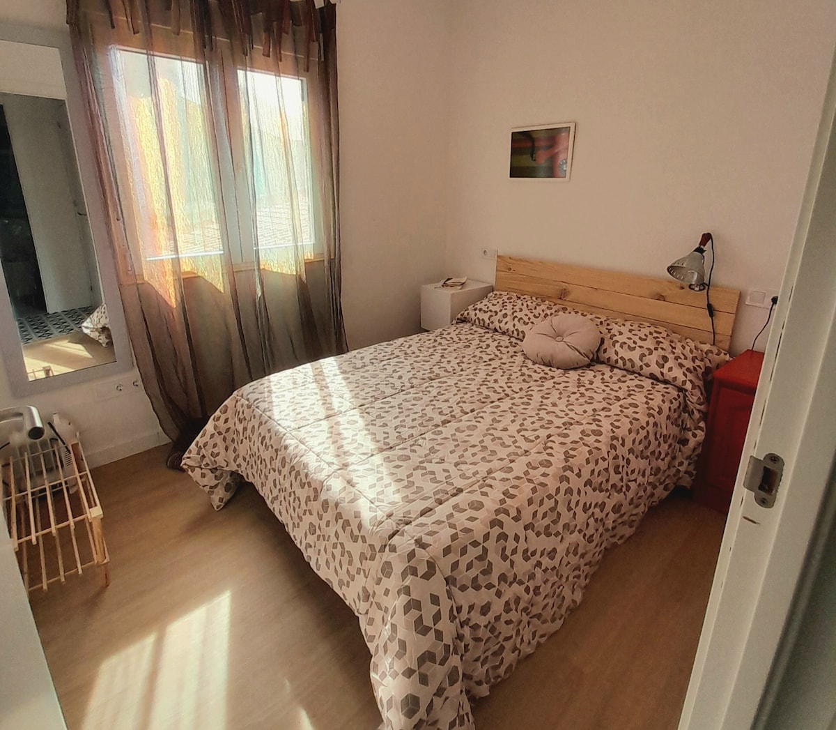 A well-lit sleeping area features a spacious bed covered with a spotted comforter. Natural light filters through sheer curtains, casting soft shadows. A bedside lamp and a small nightstand are present, with a mirror reflecting the room's inviting ambiance.