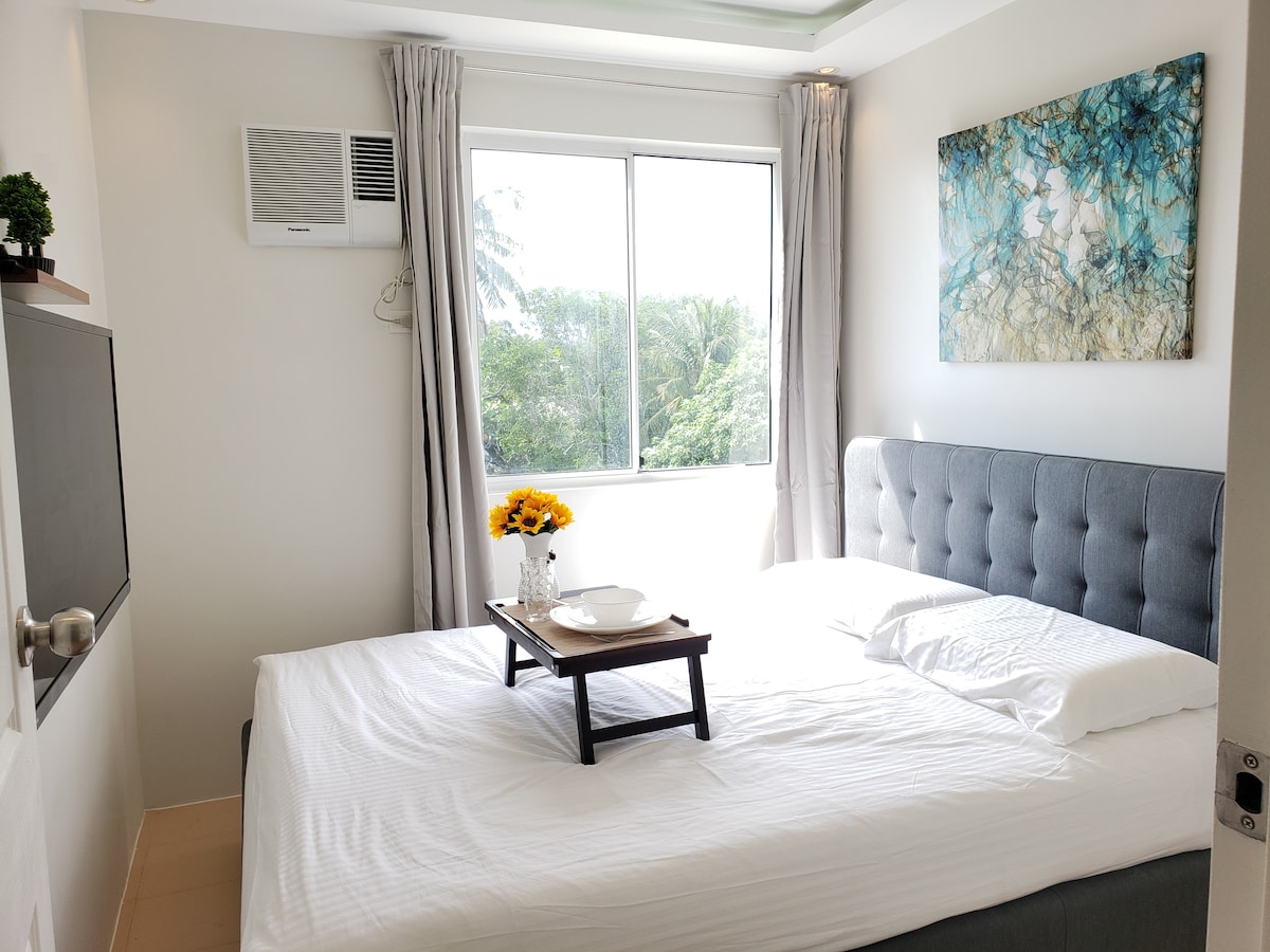 A light-filled bedroom features a spacious bed with a cozy white duvet and a tufted headboard. A wooden tray with dishes sits on the bed. A large window with sheer curtains showcases views of greenery outside. An abstract wall art piece adds a touch of color.