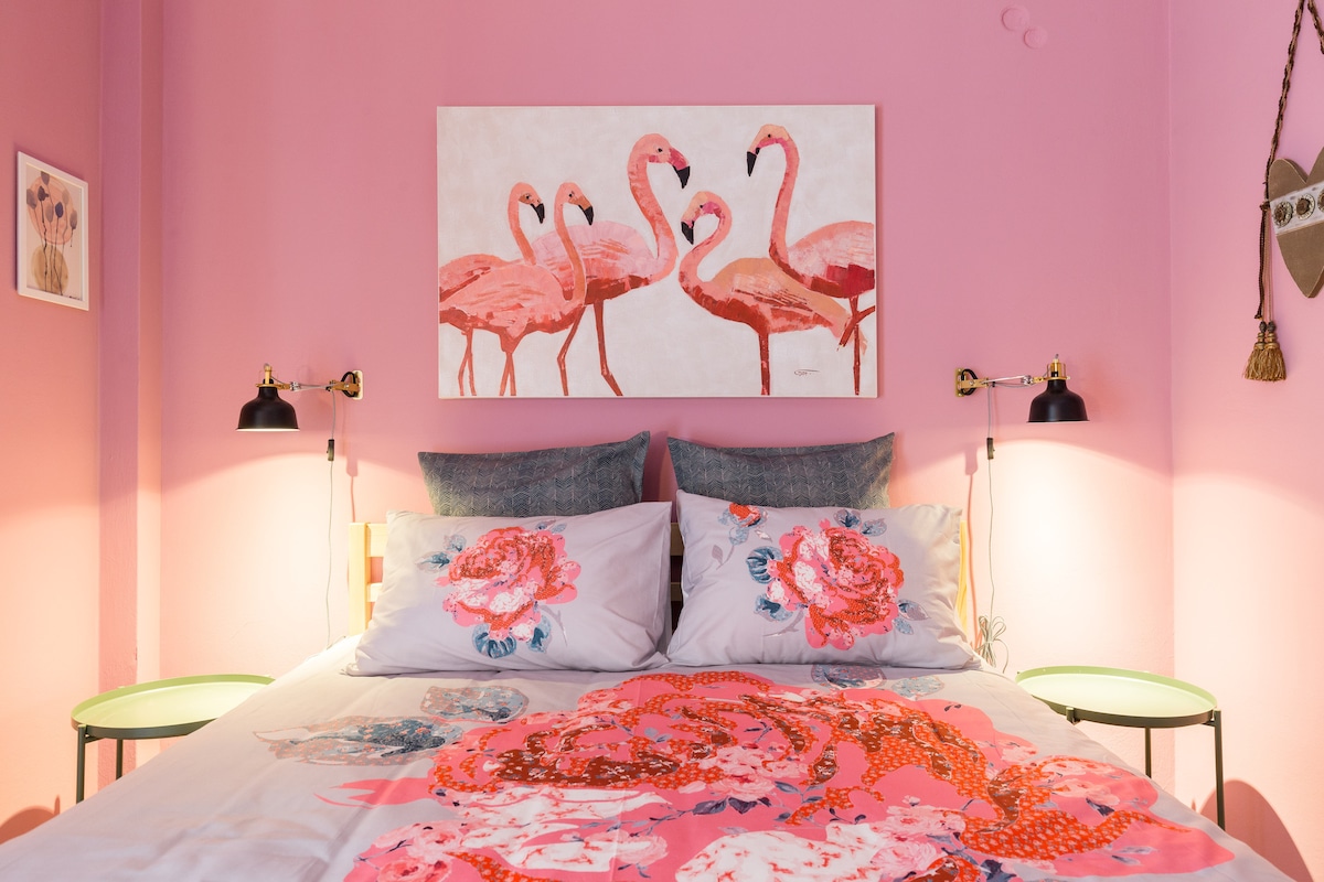The bedroom features a double bed adorned with floral bedding. On either side of the bed, green bedside tables hold modern lamps. A vibrant artwork of flamingos is displayed above the bed, complemented by soft pink walls that enhance the room's cheerful ambiance.