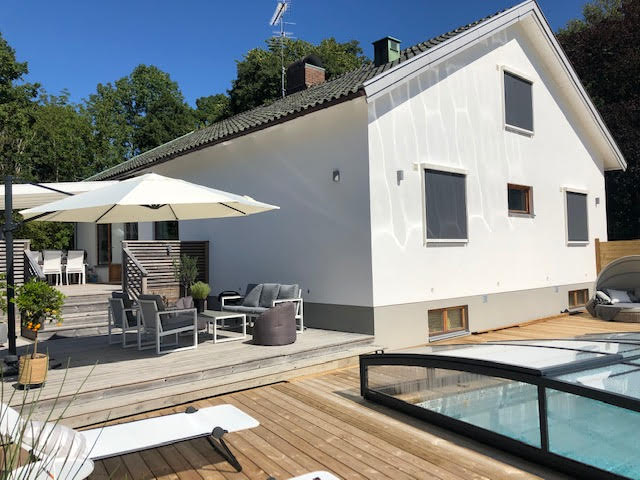 The exterior of a modern villa is presented, featuring a wooden deck with lounge seating. An umbrella provides shade over the sitting area, while a swimming pool is visible adjacent to the house. Light-colored walls and a green canopy of trees enhance the surroundings.
