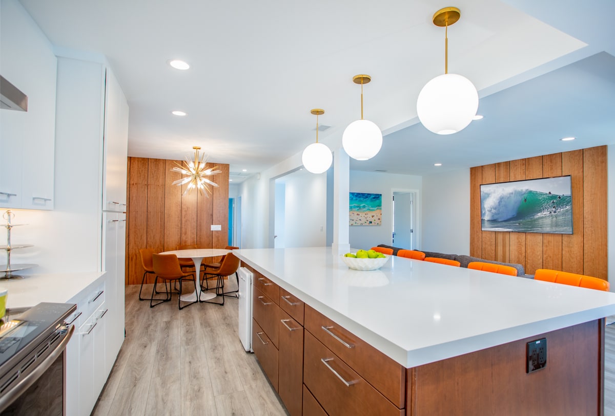 A modern kitchen features a large white island with wood cabinetry, stainless steel appliances, and stylish pendant lighting. A dining area with mid-century chairs is visible in the background, along with a lounge area showcasing colorful seating and coastal artwork on the wall.