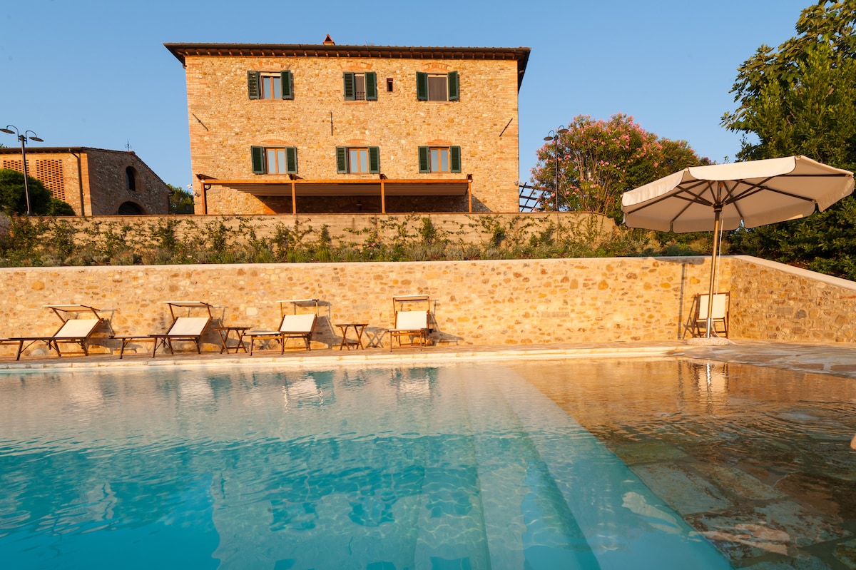 A rectangular swimming pool is situated in front of a rustic stone farmhouse. Five lounge chairs are arranged along the pool's edge, with an umbrella providing shade. The building features green shutters and is surrounded by lush landscaping.