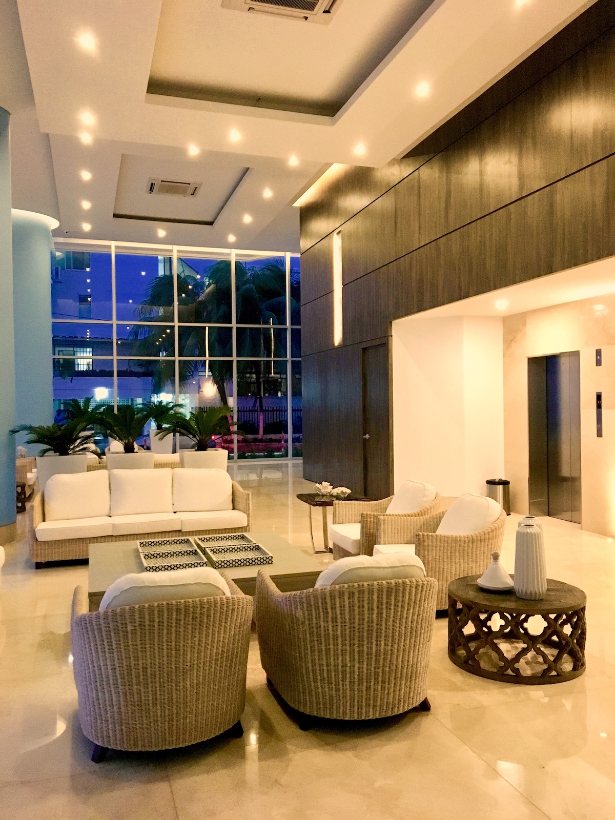 The lobby area features a modern design with large glass windows allowing natural light. A comfortable seating arrangement includes a white sofa and several armchairs. Decorative elements, such as plants and stylish lighting fixtures, create a relaxed ambiance.