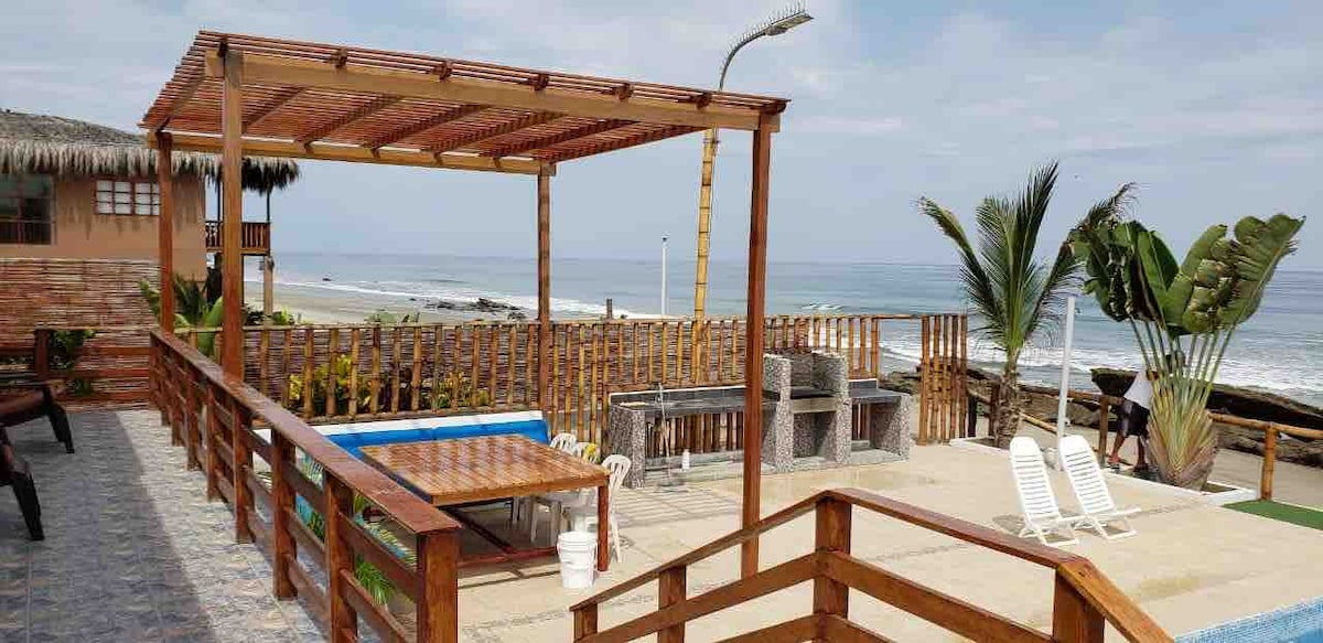 An outdoor terrace offers a shaded seating area with wooden furniture, providing views of the ocean. A swimming pool is visible next to the grill area, complemented by tropical plants. The stylish design incorporates natural materials, enhancing the connection with the seaside environment.
