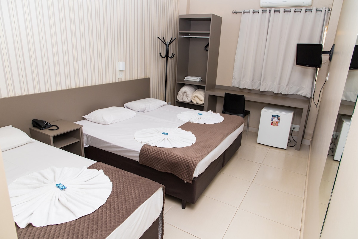 The room features two spacious beds with decorative bed covers in neutral tones. A simple workspace with a chair is positioned next to a compact closet. A wall-mounted television and a minibar are included, with light entering through a window covered by light curtains.