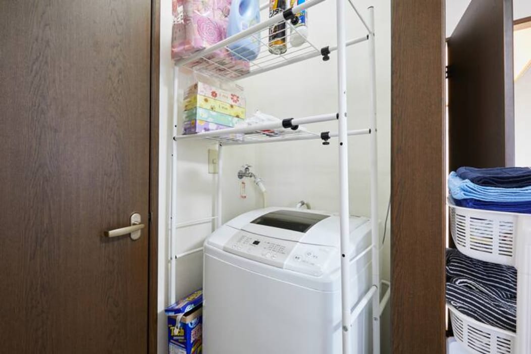 A compact laundry area is equipped with a washing machine situated beneath a multi-tiered shelving unit. Shelves hold various laundry supplies, while neatly folded towels are stored nearby in baskets. The space is enclosed by a wooden door, ensuring privacy.