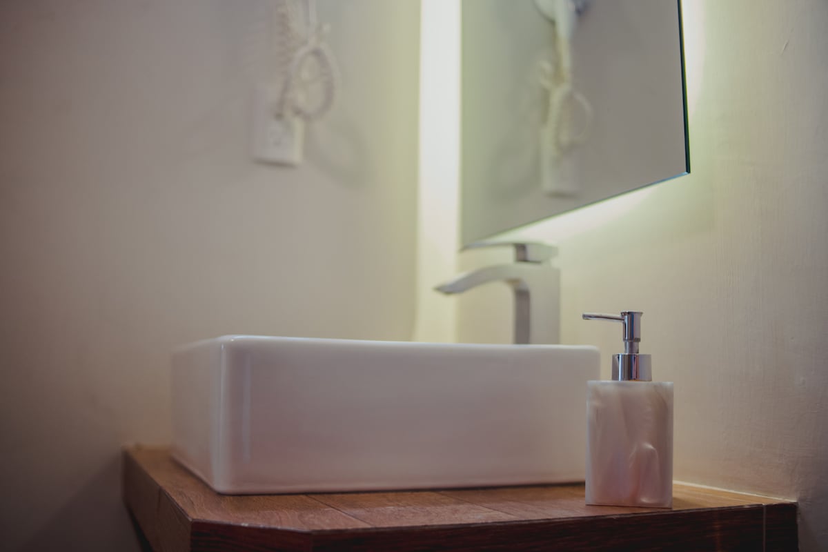 A modern bathroom countertop features a rectangular white sink paired with a sleek silver faucet. A mirrored wall hangs above, reflecting soft light. Next to the sink, a decorative soap dispenser is positioned for convenience.