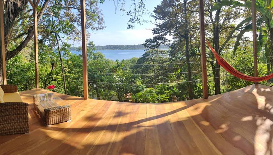 A spacious wooden deck is surrounded by lush greenery, offering a serene view of the water in the distance. A hammock is suspended nearby, and a small coffee table and chairs are positioned on the deck, inviting relaxation in nature.