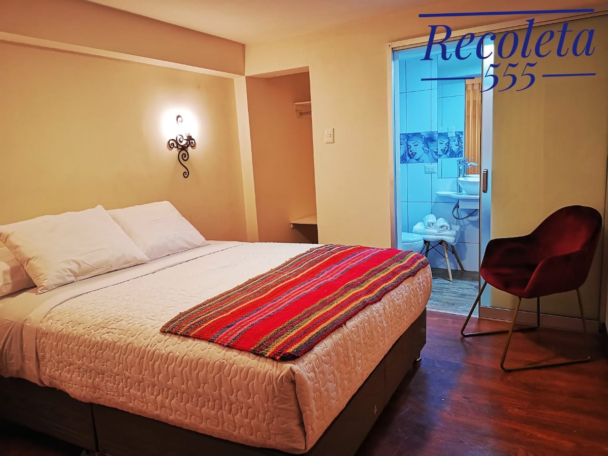 A well-furnished room features a spacious bed with white linens, complemented by a colorful striped throw. A cozy armchair in deep red adds a touch of style. A bathroom is visible in the background, showcasing tiled walls and essential amenities.
