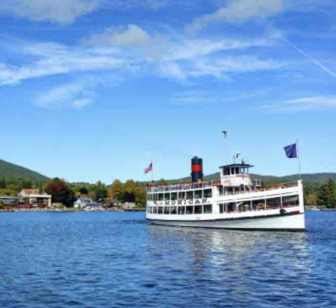 A large boat is seen navigating through calm waters, with a scenic shoreline in the background. Trees with autumn foliage surround the area, while a clear blue sky features a few clouds. The boat includes a distinct red and black smokestack.