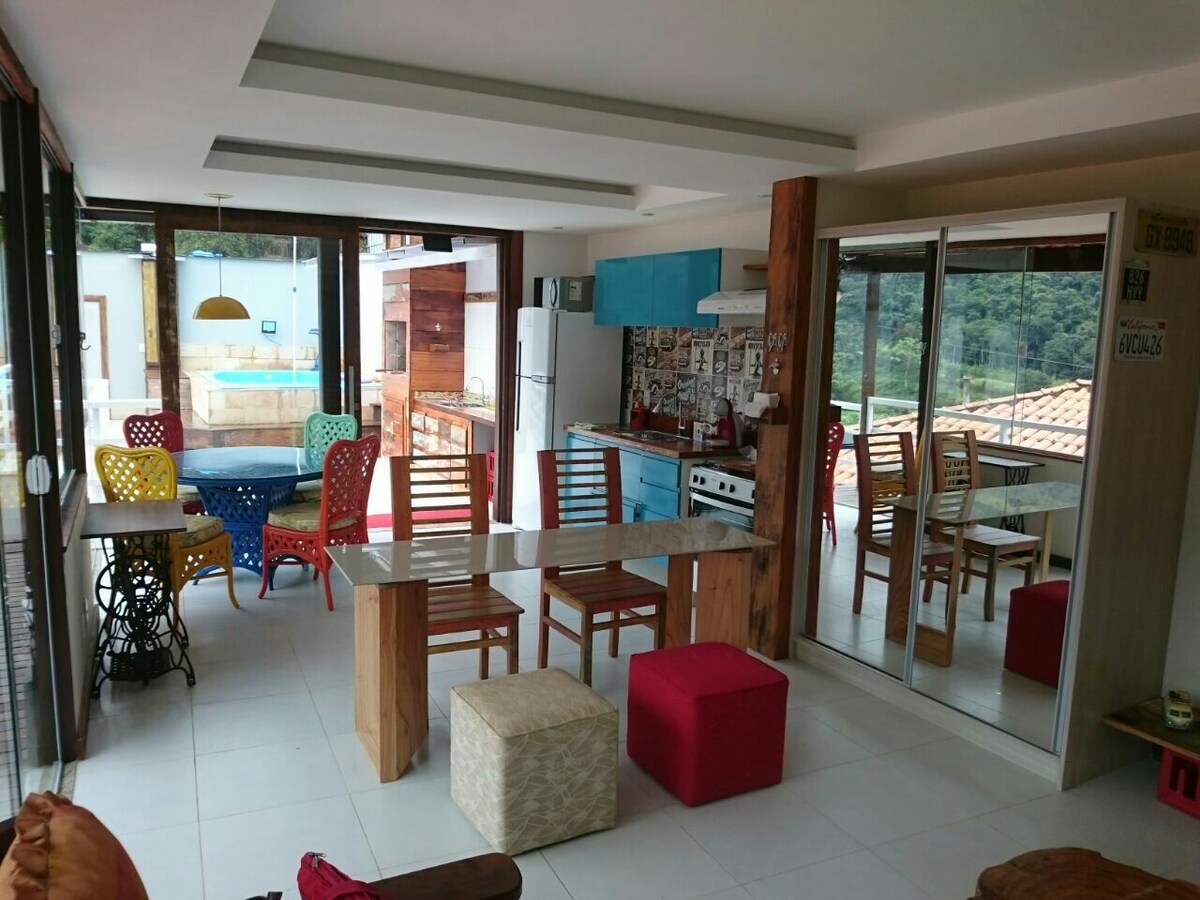 An open-plan living space features a wooden dining table surrounded by colorful chairs, with a mirrored cabinet positioned nearby. A bright kitchen is visible in the background, while large glass doors frame views of the outdoor area and pool.
