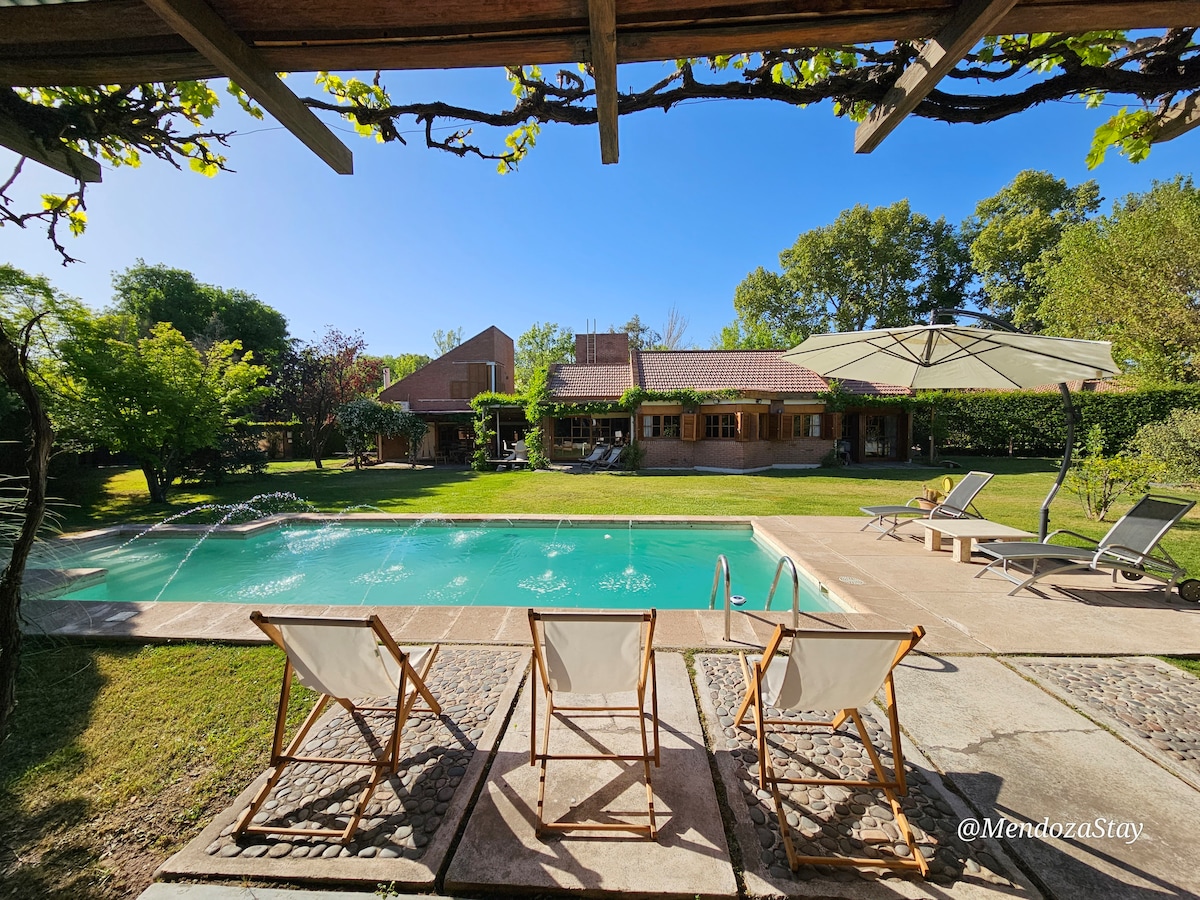 A serene outdoor area is highlighted by a clear swimming pool, surrounded by lawn and trees. Two chaise lounges face the water, while an outdoor umbrella offers shade. The house in the background features large windows and terrace access, enhancing the inviting environment.