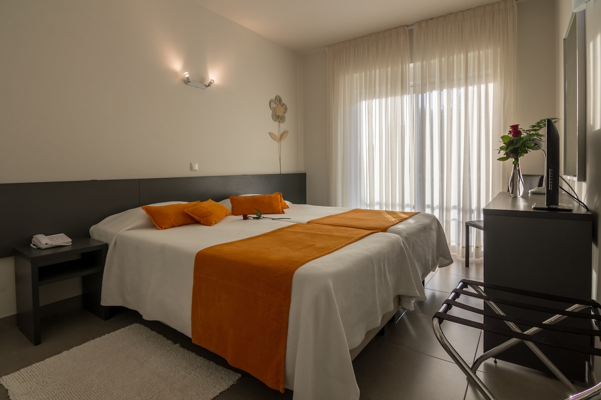 A spacious guest room is furnished with two large beds, dressed in soft linens, and accented with orange throw pillows. Natural light filters through sheer curtains, illuminating the room. A modern TV is positioned on a sleek dresser, with a small seating area featuring a folding stool.