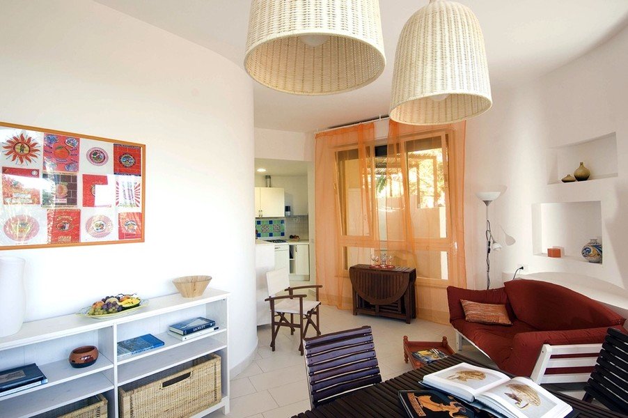 A bright living area is presented with light-toned walls and a combination of natural textures. A round table is seen at the forefront, accompanied by dark wooden chairs. A vibrant wall art piece showcases various colors, while large wicker light fixtures hang above.