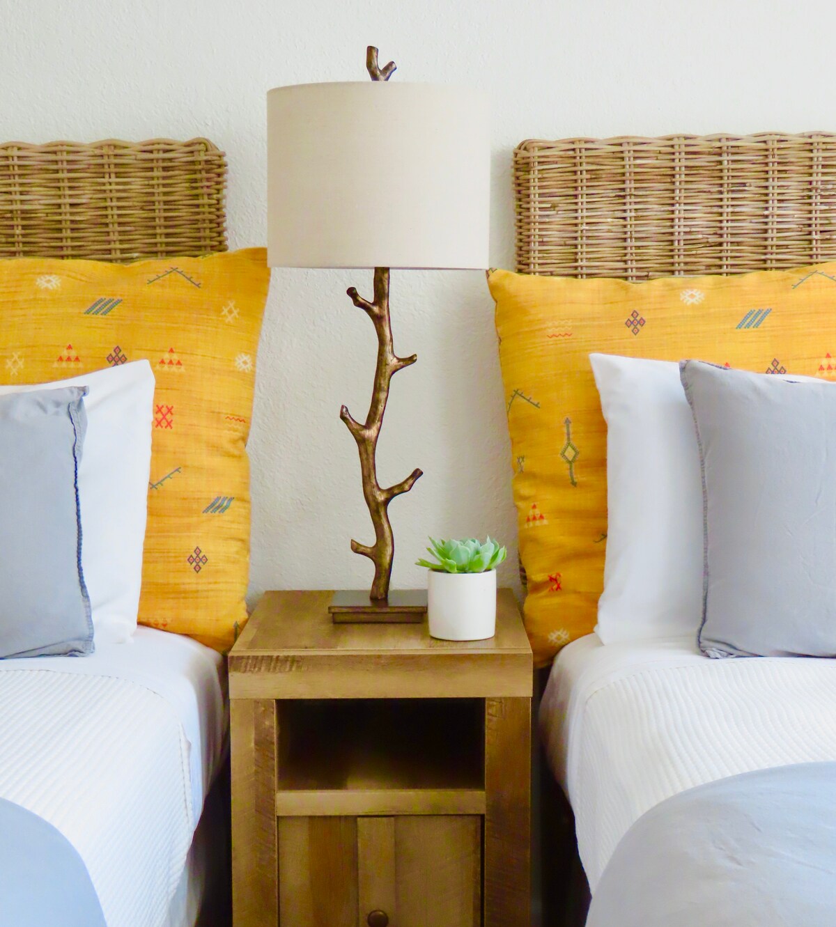 Two twin beds are positioned next to each other, each featuring decorative yellow-patterned headboards. A wooden nightstand is placed between them, topped with a branch-shaped lamp and a small potted plant. Soft gray pillows add contrast to the bedding.