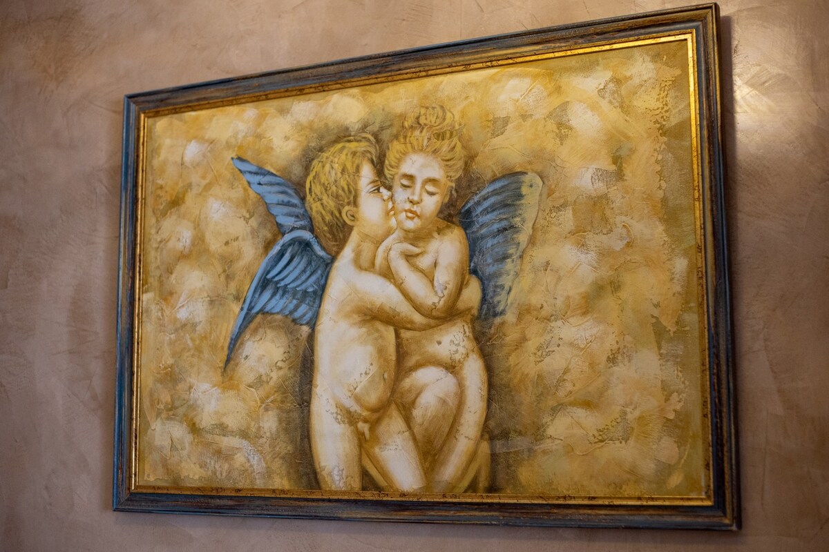A framed artwork is displayed on a textured wall, depicting cherubic figures with blue wings embracing each other. The soft, muted color palette adds warmth to the scene, while the intricate details of the figures are gently highlighted by the lighting.
