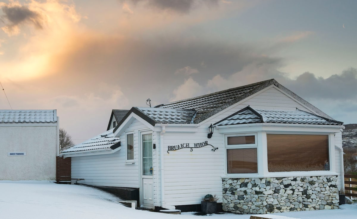 The exterior of the cottage is visible, showcasing a modern design with large windows. Snow blankets the ground, highlighting the structure's light-colored siding and stone accents. A cloudy sky suggests a serene atmosphere, with soft sunlight peeking through, adding warmth to the scene.