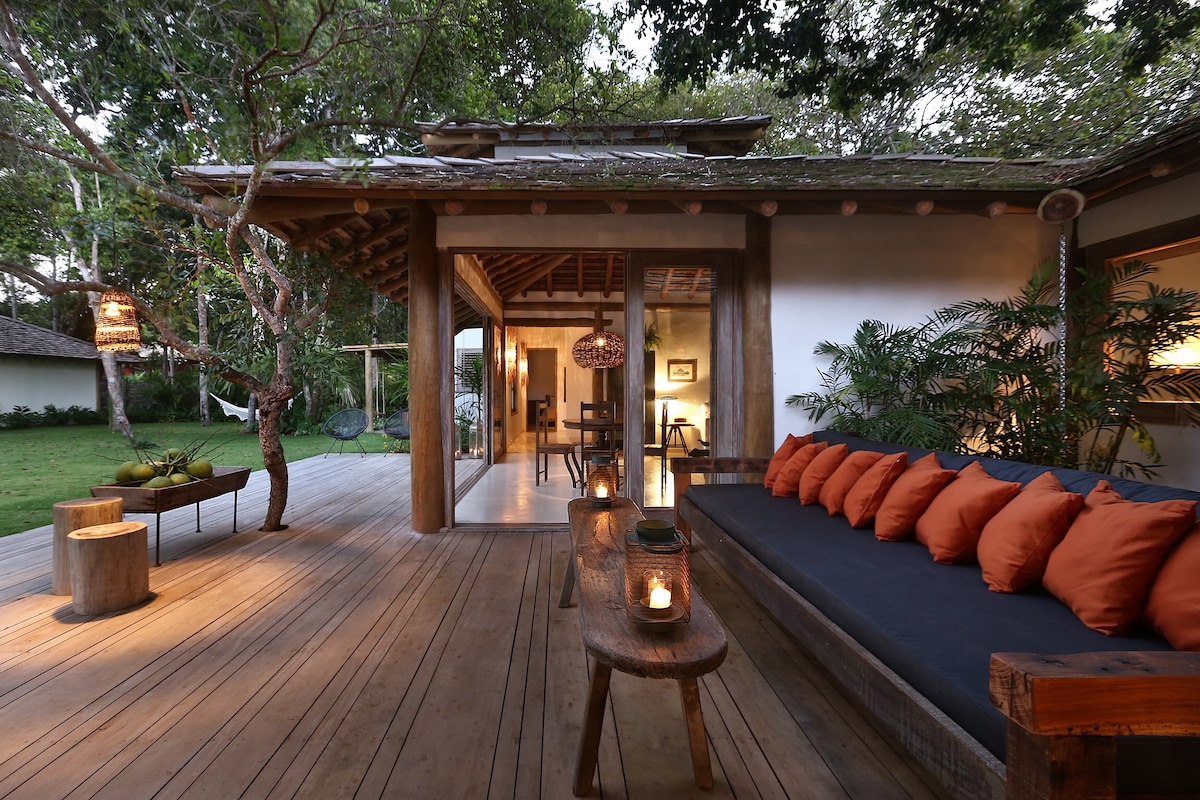 An outdoor seating area is featured on a wooden deck, furnished with orange cushions on a dark sofa. A wooden table holds candles, creating a warm ambiance. Tropical plants and trees surround the space, enhancing the connection to nature.