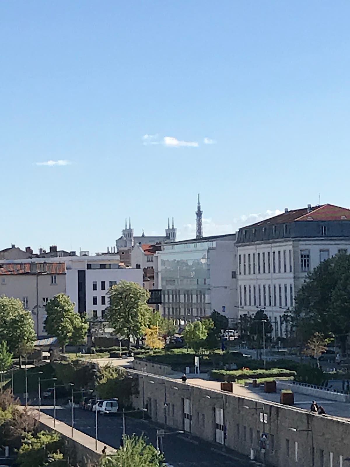 The image captures a cityscape view featuring buildings of varying architectural styles, with greenery in the foreground. Notable structures include spires and a distant tower, while the sky is clear with a few scattered clouds, enhancing the bright atmosphere.