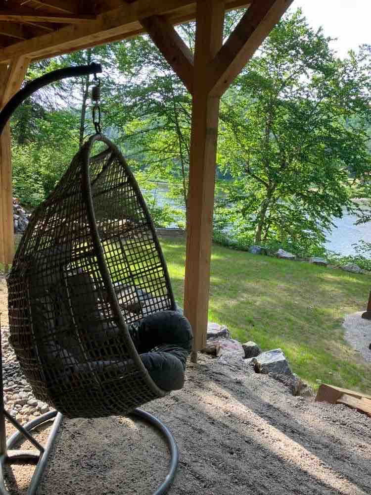 A woven hanging chair is suspended from a wooden structure, providing a comfortable spot to relax. Surrounding greenery and a glimpse of the river enhance the natural setting. The ground is covered with smooth gravel, creating a serene atmosphere.