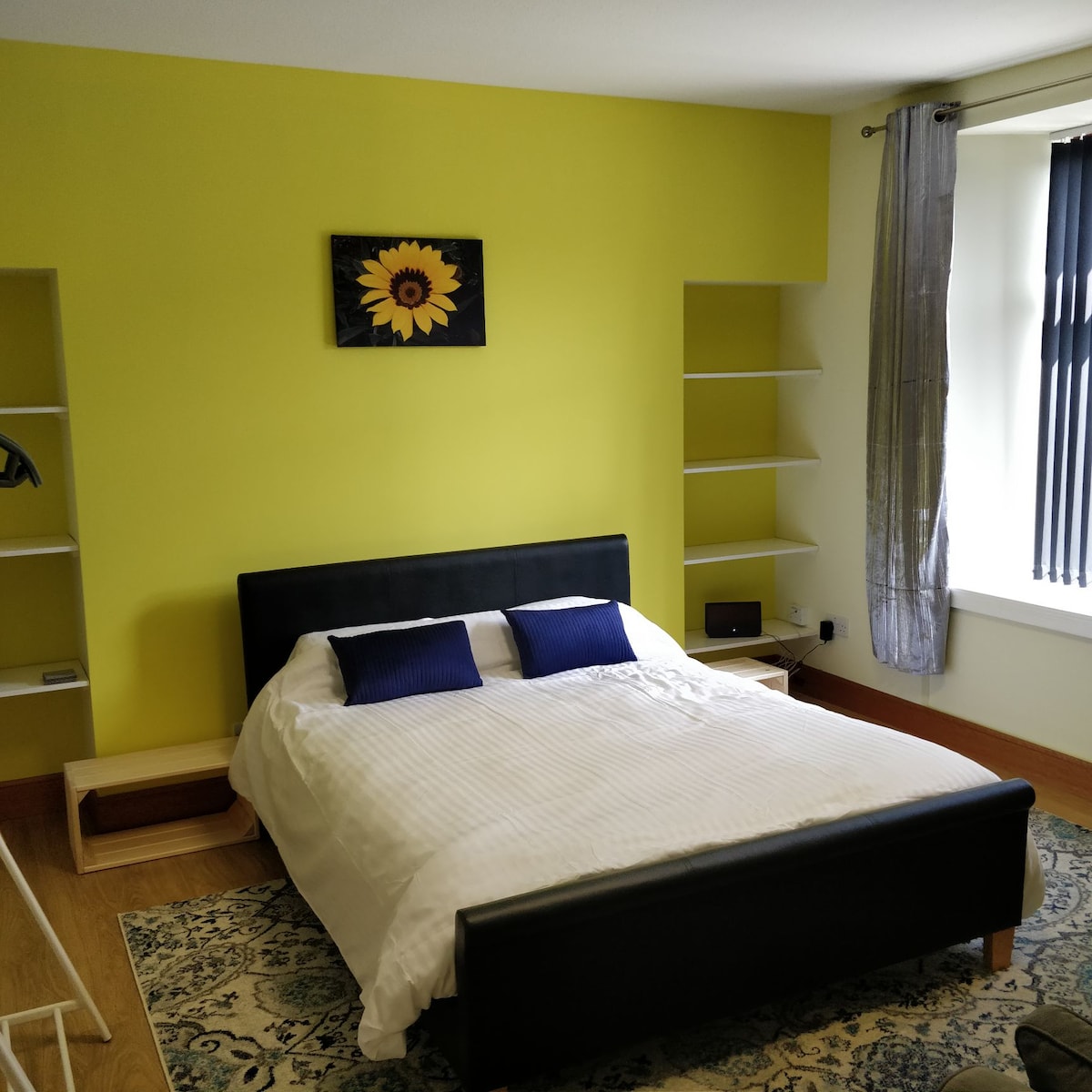 A bright bedroom features a sunlit yellow wall and a spacious bed dressed in white linens with two blue accent pillows. A small bedside table sits next to the bed, and a sunflower artwork adorns the wall above. Shelves are provided for additional storage.