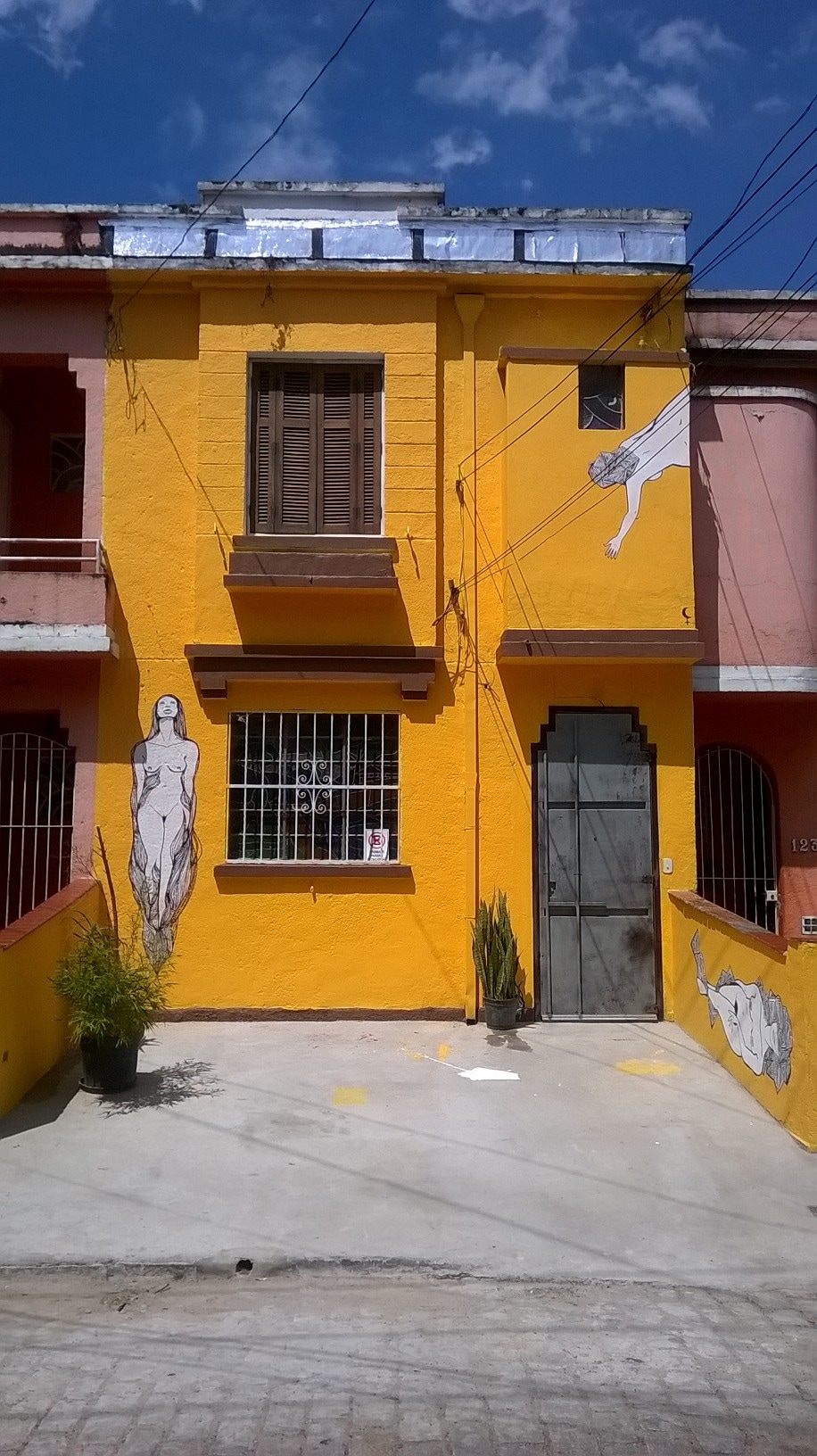 The exterior of a vibrant yellow building is featured, adorned with artistic murals of human figures. Tall windows with wooden shutters complement the entrance, surrounded by greenery. A concrete pathway leads to the door, framed by a colorful wall, under a clear blue sky.