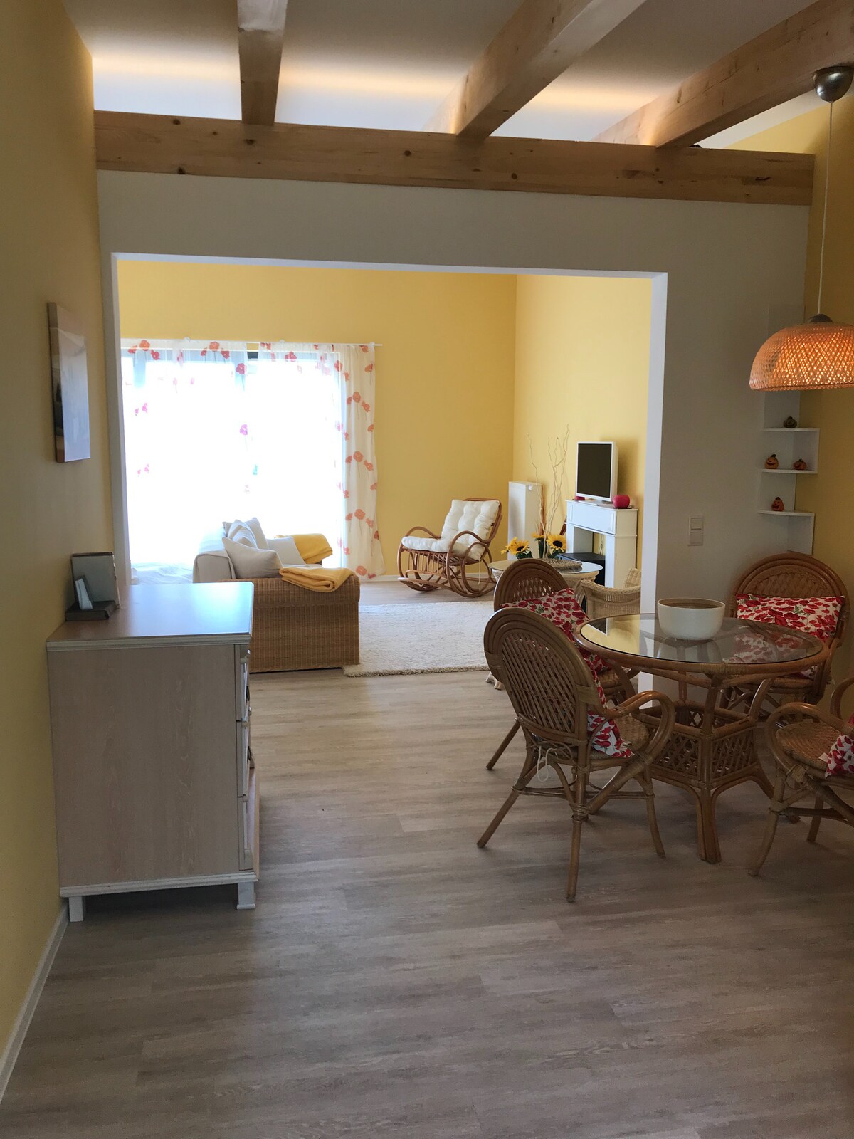 An open layout connects a dining area with a round table surrounded by rattan chairs to a bright living space. Light pours through large windows adorned with colorful curtains, illuminating a cozy seating arrangement with two cushioned chairs and a small TV stand.