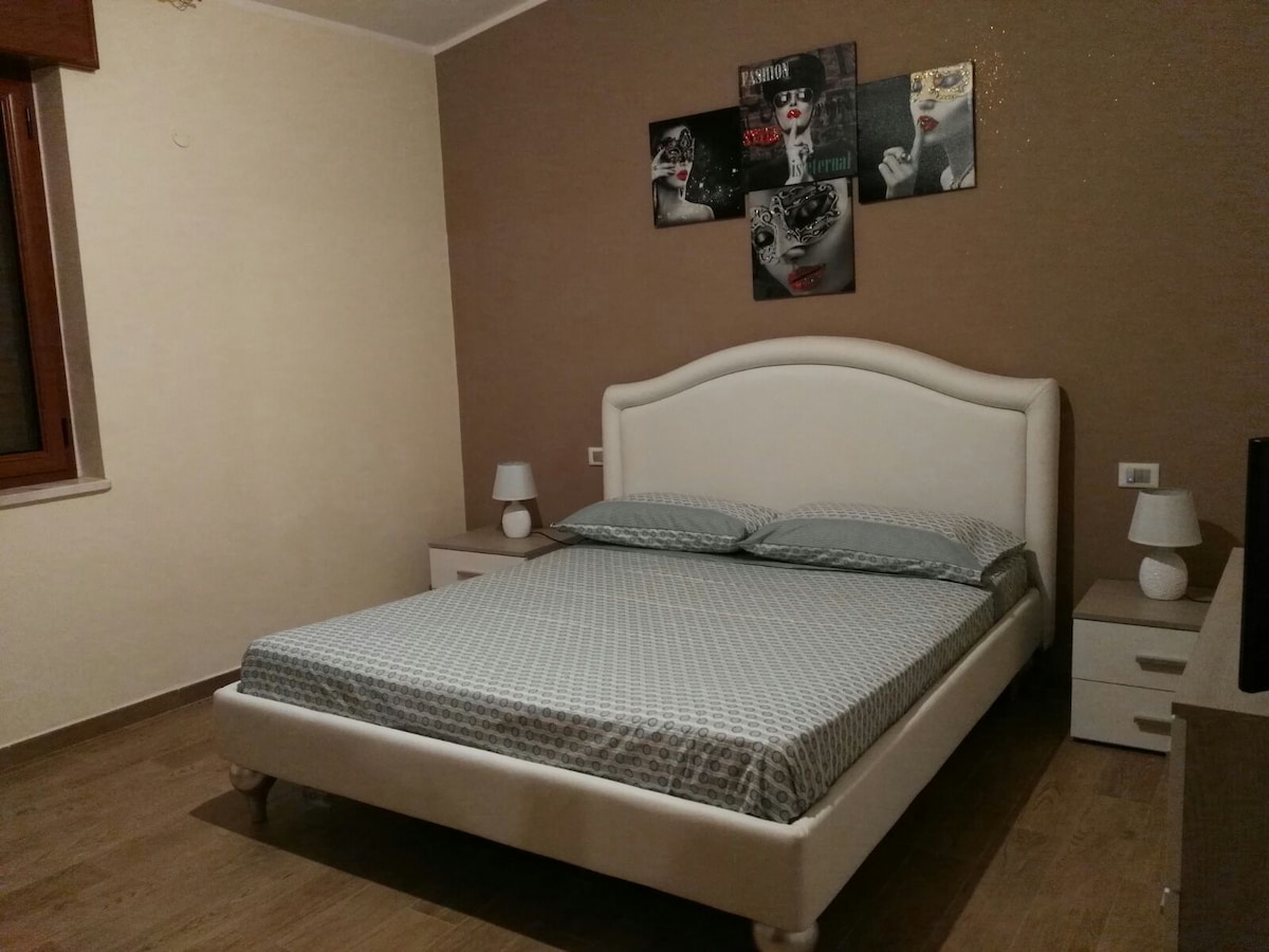 A spacious bedroom is presented with a soft upholstered bed featuring a light-colored frame. The bed is dressed in a patterned blue and white coverlet. Two bedside tables with lamps are positioned on either side, and art decor is displayed on the wall above.