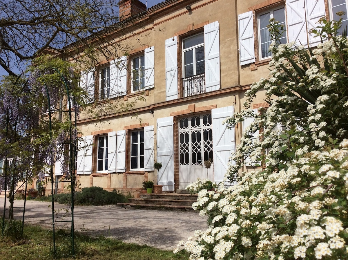 An elegant two-story building features multiple white-shuttered windows and a central door framed by decorative wrought iron. A lush garden with blooming white flowers and greenery enhances the welcoming exterior, while steps lead up to the entrance.