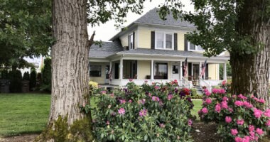A charming two-story house is framed by lush green trees and colorful flowering bushes. The exterior features yellow siding, a welcoming porch with seating, and American flags. A manicured lawn extends in the foreground, enhancing the inviting appearance of the property.