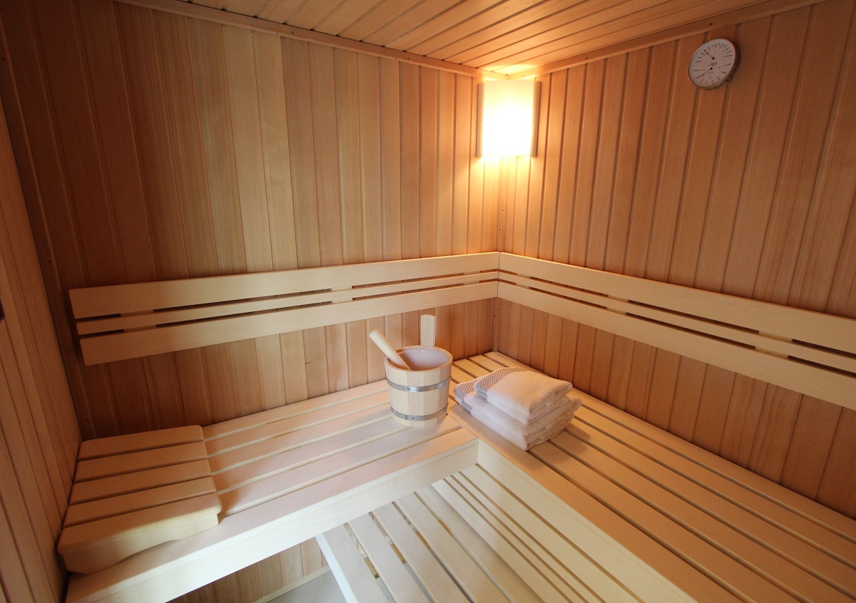 A wooden sauna is featured, showcasing light-colored wooden benches arranged in a corner layout. A small clock is mounted on the wall, and a bucket along with towels is placed on the seating. Warm lighting creates a calming atmosphere.