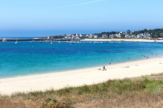 A serene beach scene showcases soft, white sands meeting clear turquoise waters. Several boats can be seen anchored in the distance, with individuals enjoying leisurely activities along the shore. A calm blue sky stretches above, enhancing the tranquil atmosphere of the coastal landscape.
