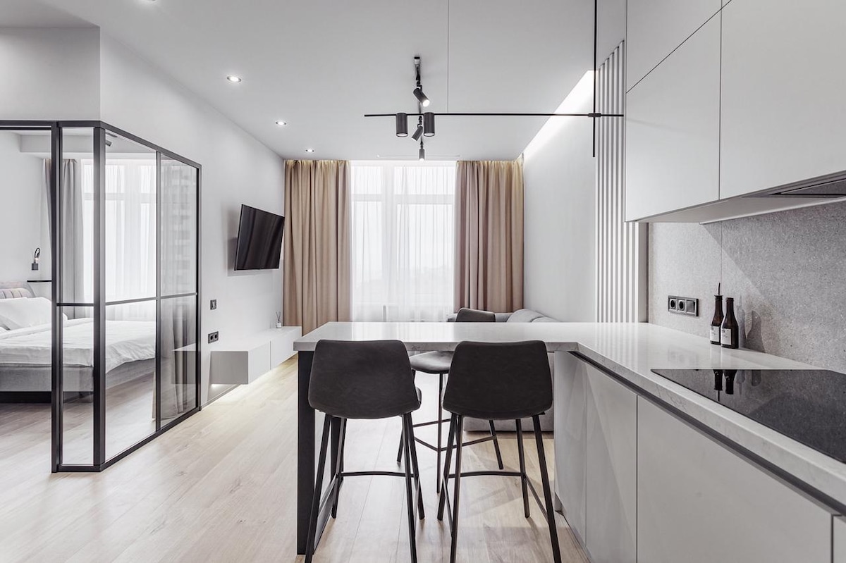 An open-plan living area features a sleek kitchen island with two high stools and modern appliances. Soft curtains frame a large window, allowing natural light to fill the space. Opposite, a wall-mounted TV is visible, with a cozy seating area partially seen in the background.