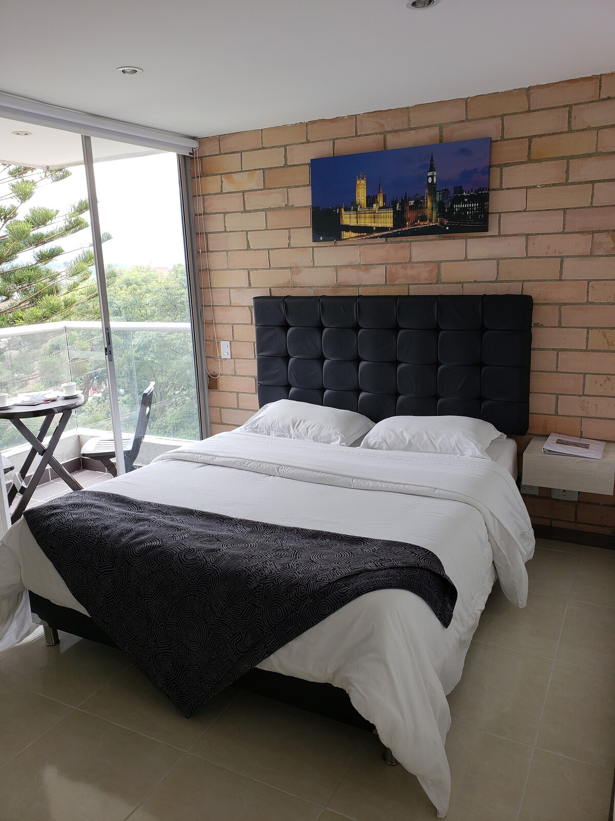 A well-appointed bedroom features a black tufted headboard above a spacious bed covered with white linens. A grey patterned blanket is draped across the foot of the bed. Natural light streams in through large glass doors that lead to a balcony with a small table and chair set.