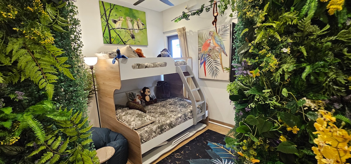 A cozy bunk bed with a plush mattress is positioned against a backdrop of vibrant greenery and artwork. The space features decorative plants, a lamp providing soft light, and colorful wall art depicting tropical scenes, creating a serene and inviting atmosphere.