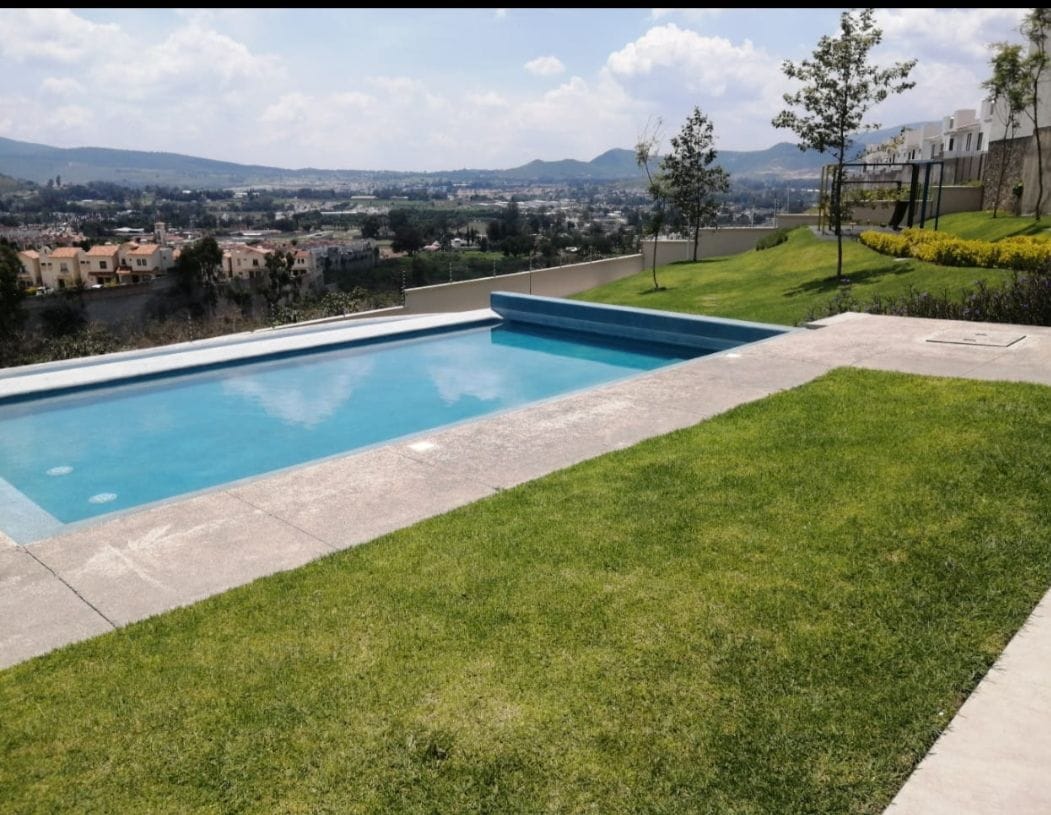 A serene outdoor area features a glistening pool bordered by smooth stone tiles, with lush green grass extending into the landscape. The distant hills and urban view create a peaceful backdrop under a clear sky.