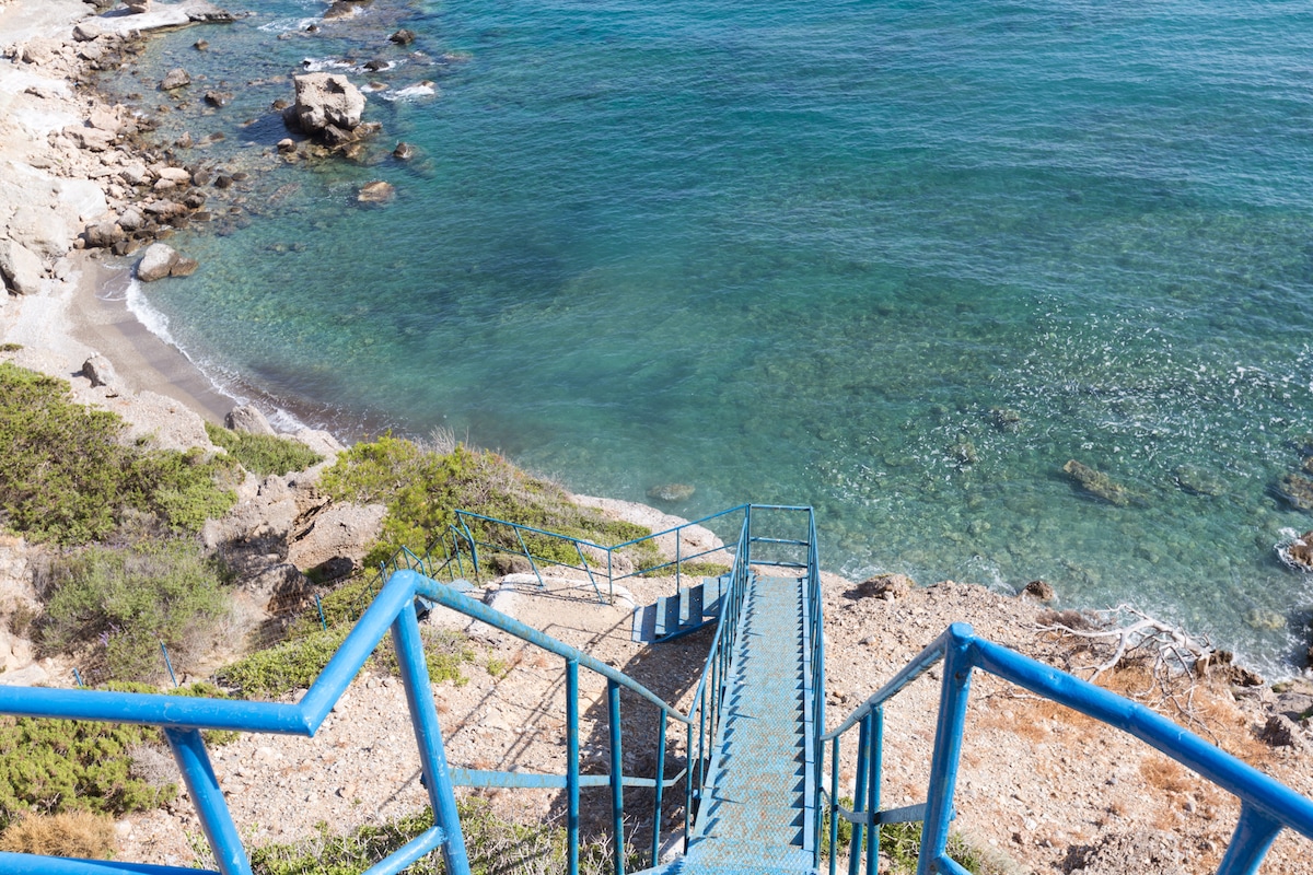 A blue metal staircase descends toward a rocky coastline, leading to clear turquoise waters. The surrounding area features patches of greenery and rocky outcrops, creating a natural coastal landscape. Sunlight reflects off the water, enhancing the scene's serene quality.