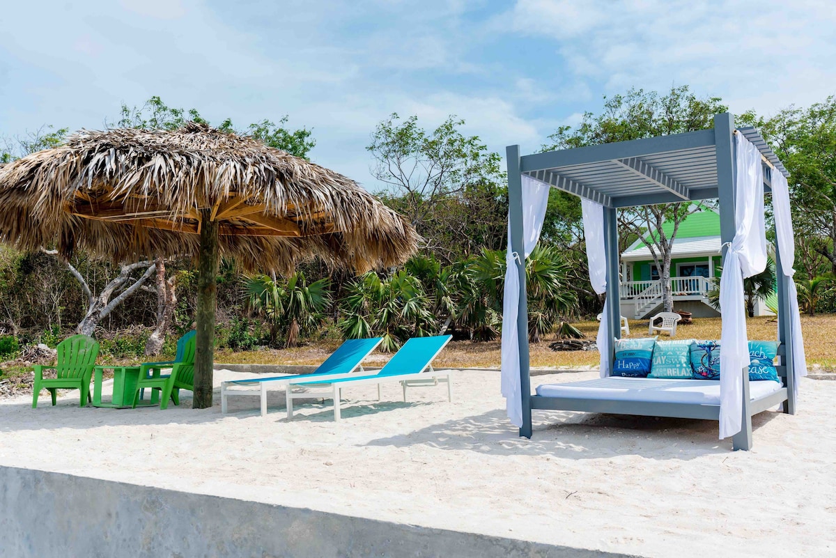 A man-made beach area features a thatched-roof umbrella shade beside a daybed with light fabric draping. Two lounge chairs in vibrant blue are positioned nearby on the sandy surface, inviting relaxation. Lush greenery and a distant cottage are visible in the background.