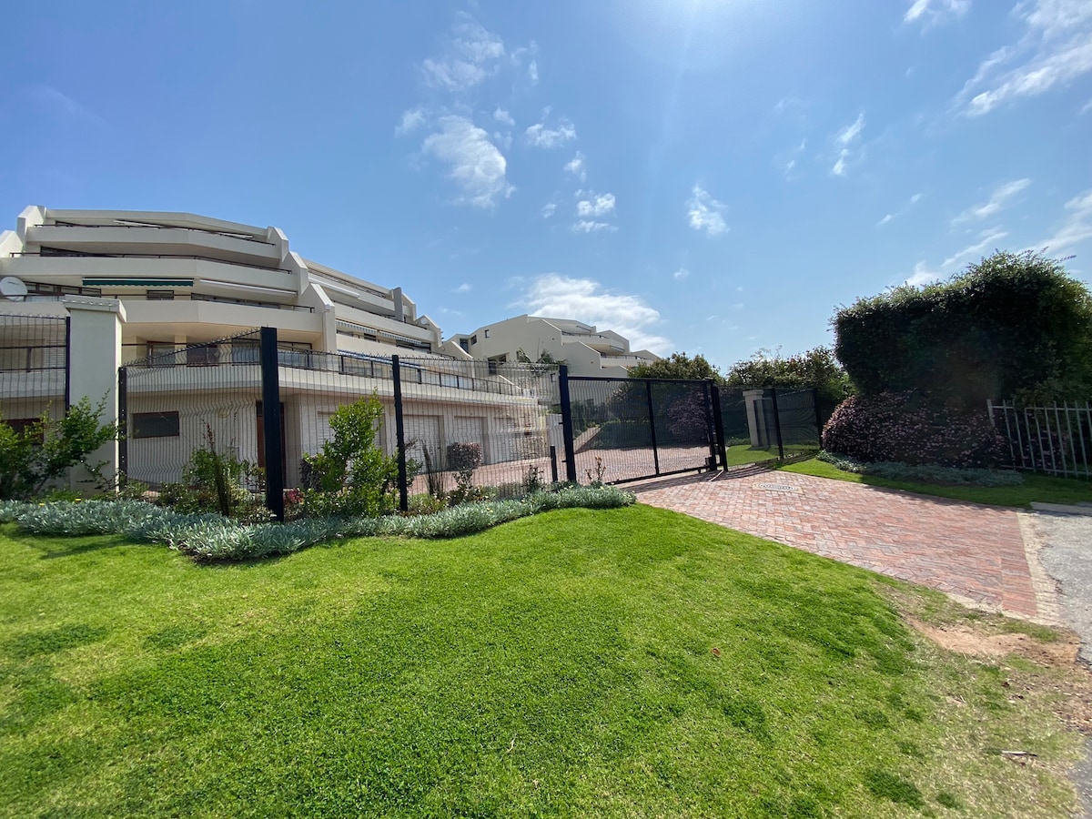 A gated entrance is visible, featuring a well-maintained lawn with lush green grass and decorative bushes. The surrounding buildings provide a modern architectural backdrop, while a clear blue sky enhances the outdoor ambiance.