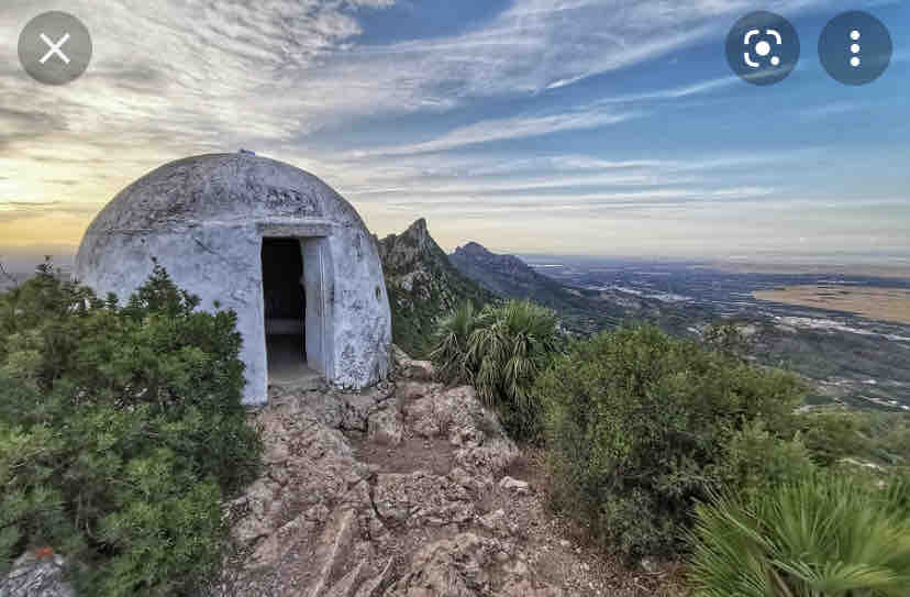 A dome-shaped structure is positioned on rocky ground, surrounded by sparse greenery. Rolling hills and mountains can be seen in the distance, offering a panoramic view of the landscape below. The sky features soft clouds and hints of color from the sunset.