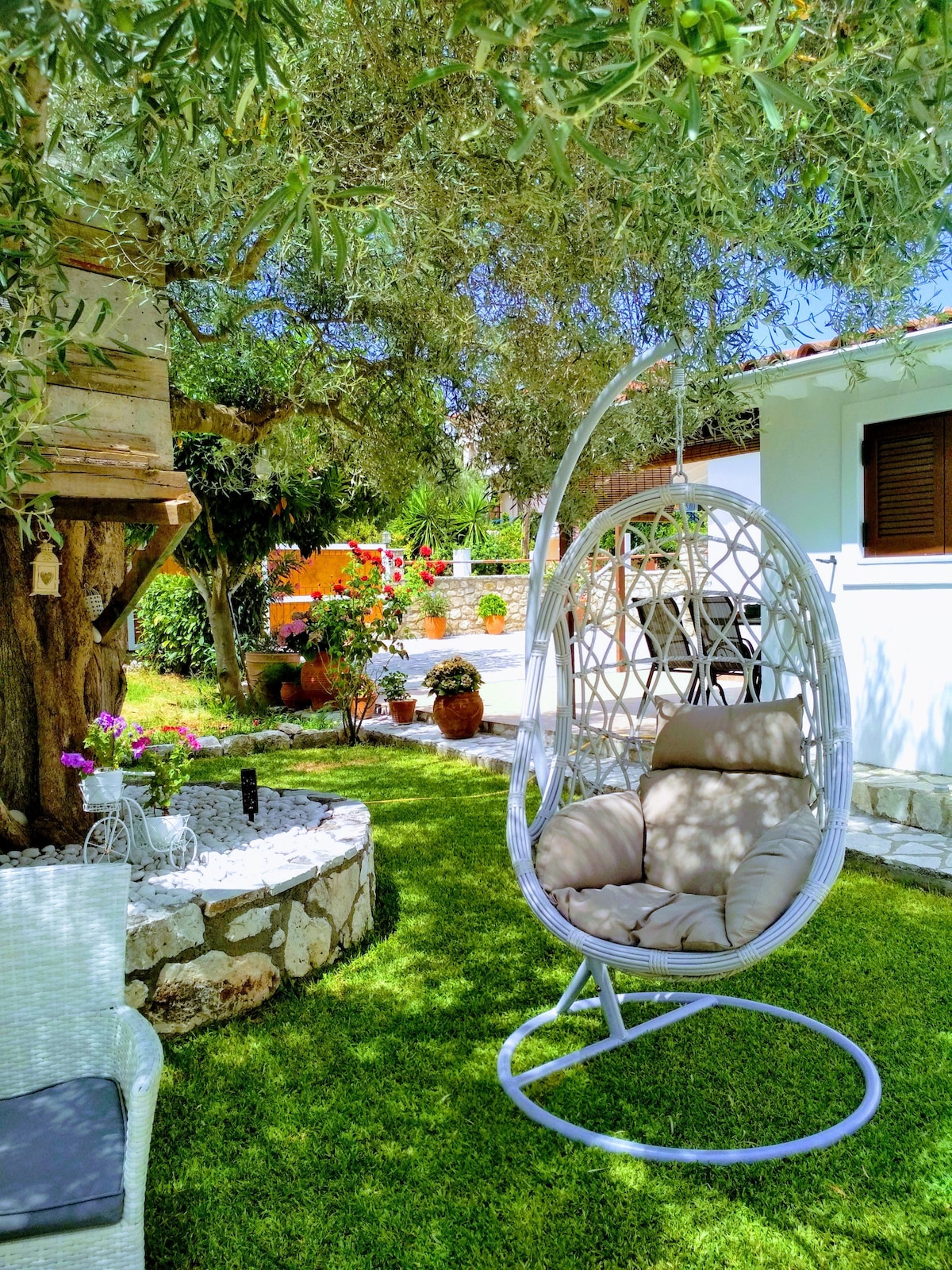 A hanging chair is positioned on a grassy area, surrounded by lush greenery and colorful flower pots. In the background, a tree house and a well can be seen, while a well-maintained patio area is visible, providing an inviting outdoor space for relaxation.
