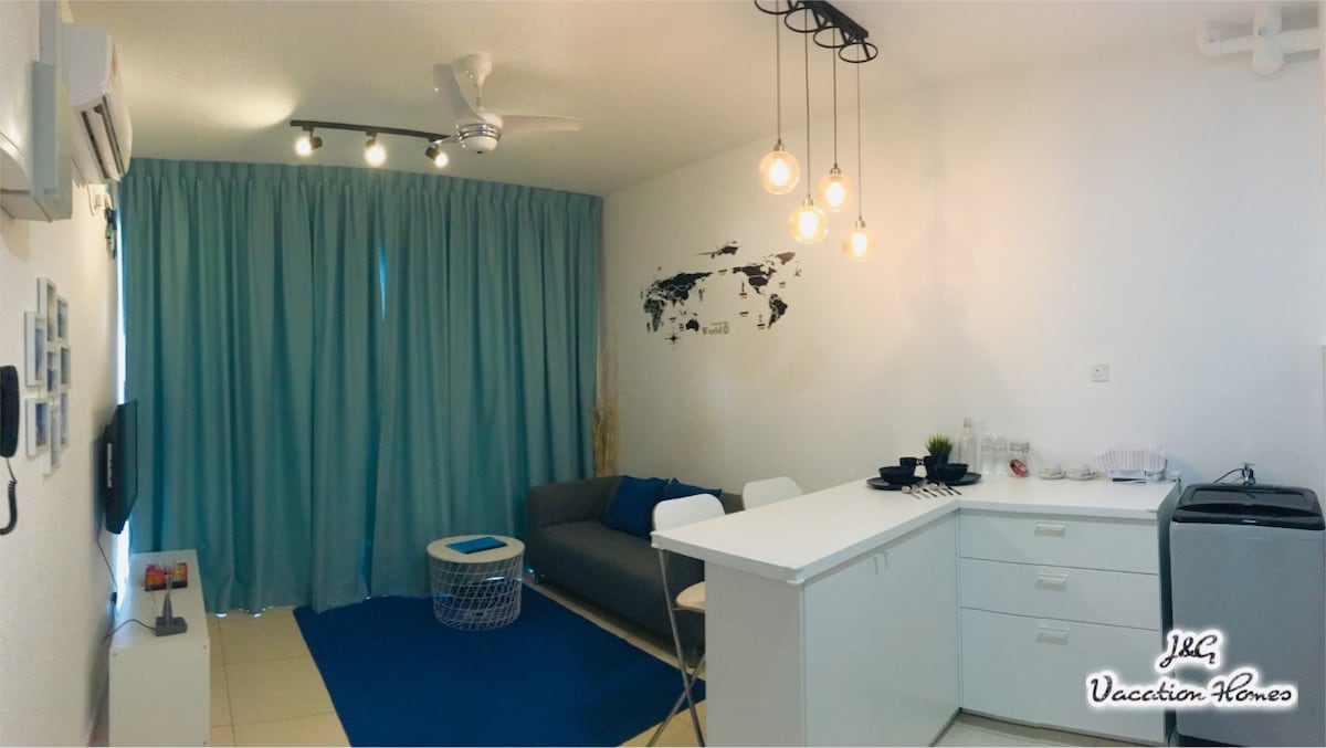 A bright living area features a gray sofa and a white coffee table. A small kitchen counter with high stools is adjacent, while soft blue curtains frame a window. Three pendant lights hang above, adding warm illumination to the space.