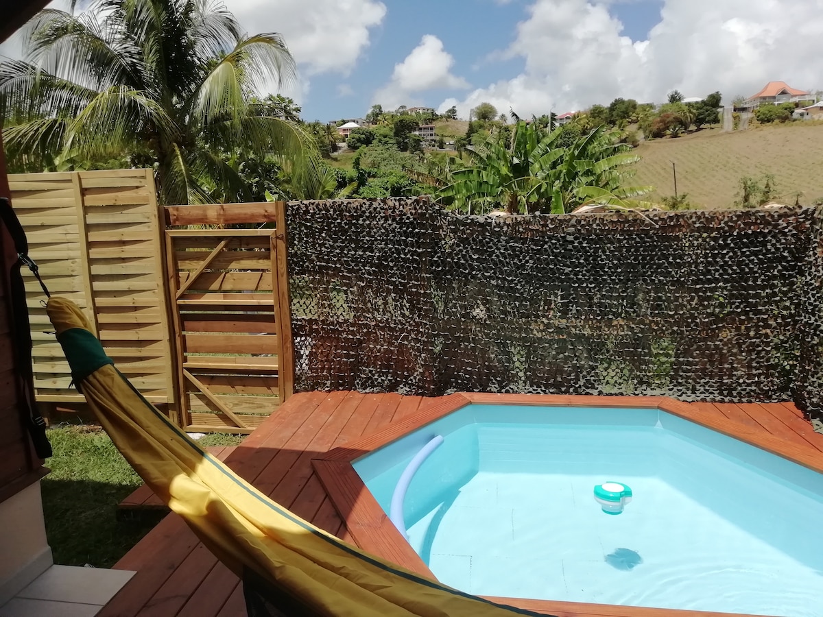 A private pool is surrounded by wooden decking and a discreet fence, providing privacy. A vibrant hammock is visible in the foreground, inviting relaxation. Lush greenery and hills are seen in the background under a partly cloudy sky.