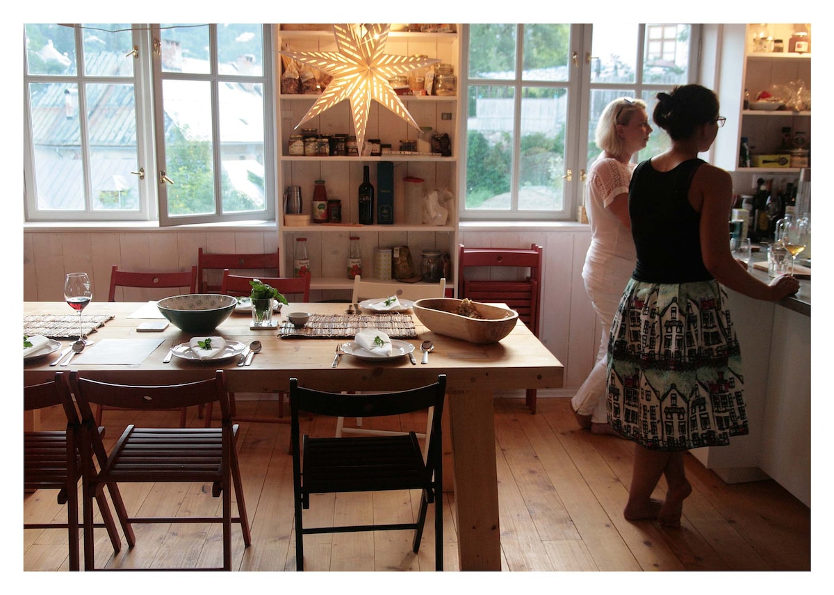 A well-lit dining area is equipped with a large wooden table surrounded by folding chairs, set for a meal. Shelves filled with kitchen items and greenery are visible. Large windows allow natural light to fill the space while offering views of the surroundings.
