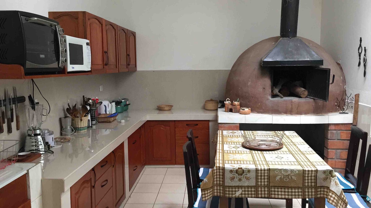 A spacious kitchen is presented, featuring wooden cabinetry and modern appliances, including a microwave. A large stone oven is situated against one wall, accompanied by a dining table set with a patterned tablecloth. Various cooking utensils and tools are organized along the counter.