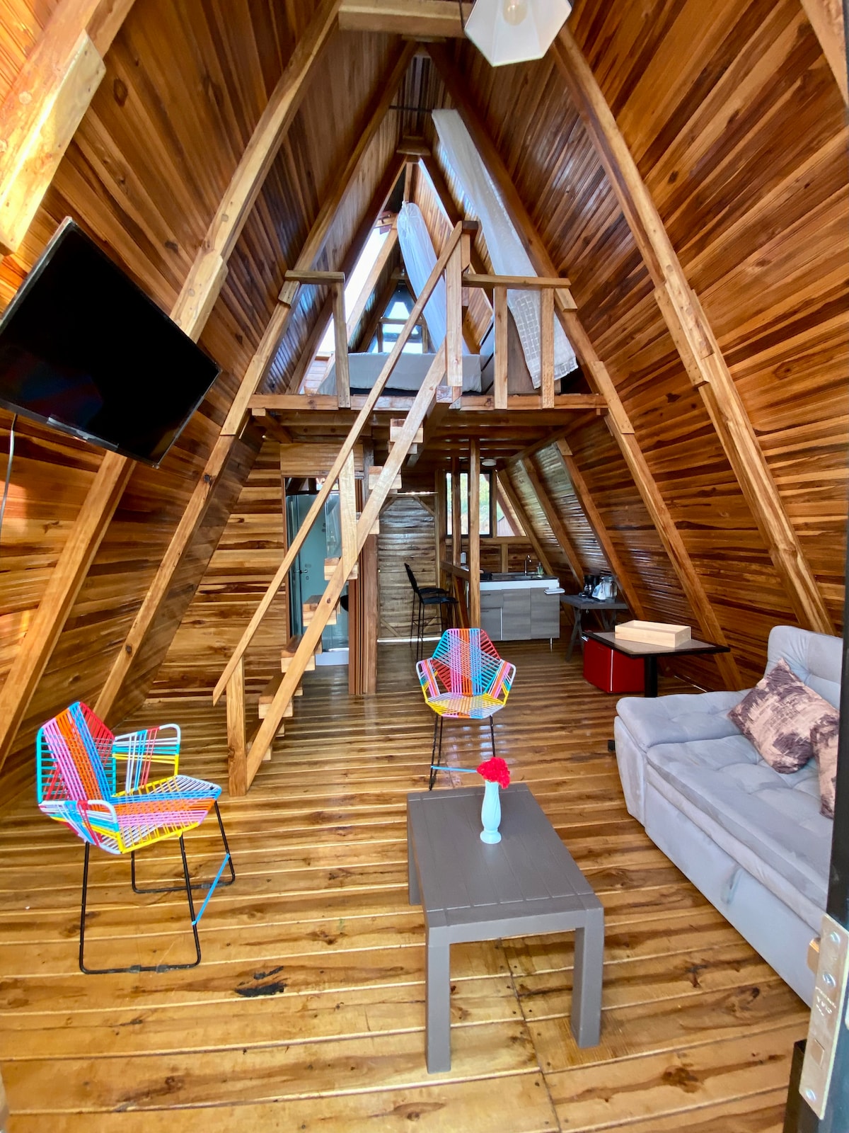 An interior space features wooden walls and a high, angled ceiling. A comfortable gray sofa faces a small coffee table, while colorful chairs are positioned nearby. A staircase leads to an upper level, and a television is mounted on the wall.